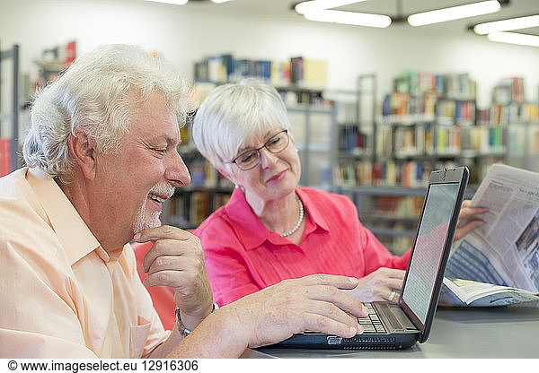 Senior couple with laptop and newspaper in a city library