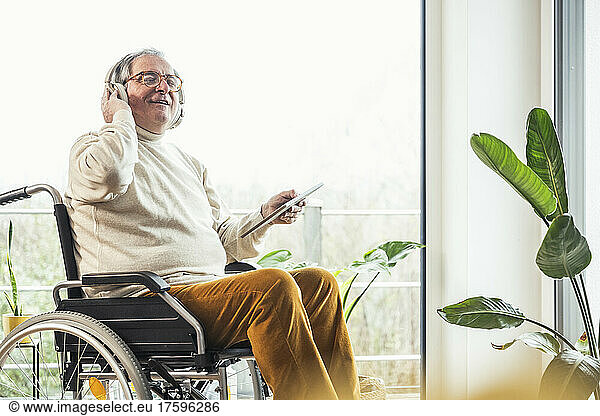 Senior disabled man with eyes closed listening music through wireless headphones on wheelchair at home