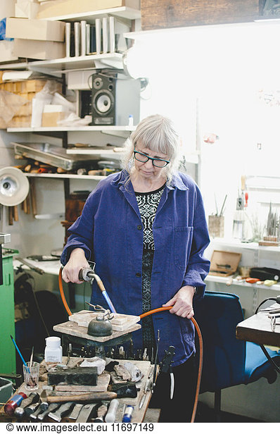 Senior female craftsperson using blowtorch for metalworking in jewelry workshop