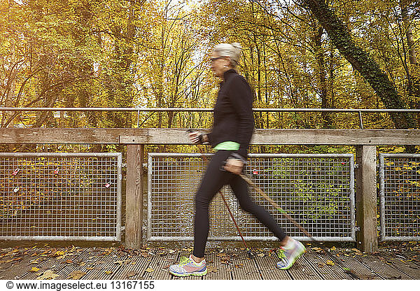 Senior female nordic walker striding over park footbridge