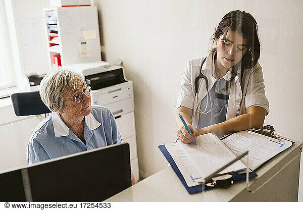 Senior female nurse looking at doctor writing prescription at medical clinic