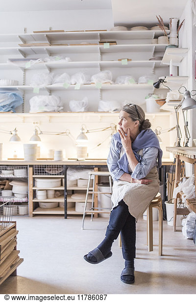 Senior female potter looking away while sitting on stool against shelves in workshop
