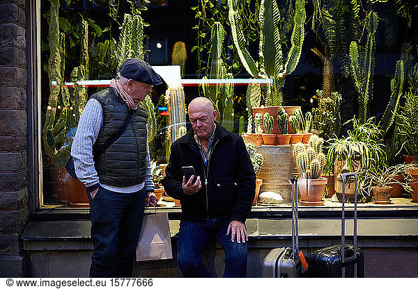 Senior gay couple booking taxi through mobile app while waiting outside plant store at night
