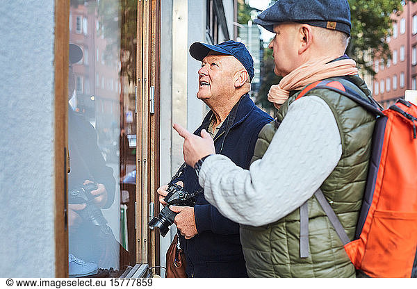 Senior gay couple standing by store window with camera in city during vacation