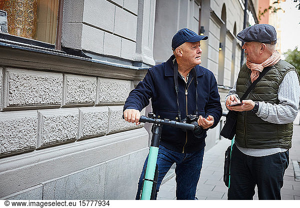 Senior gay couple using push scooters while holding mobile phone in city