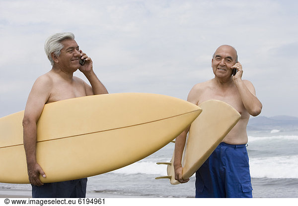 Senior Hispanic men holding surfboards on beach and talking on cell phones