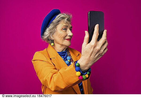 Senior influencer taking selfie using smart phone against pink background