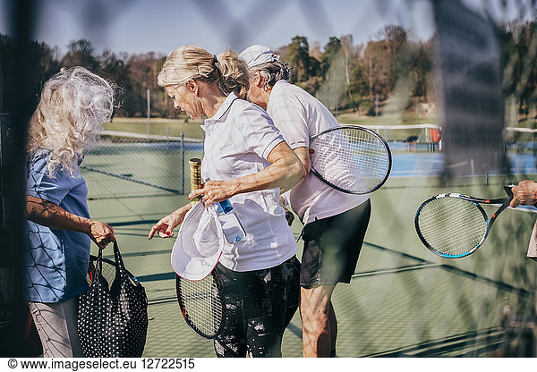 Senior male and female friends with rackets and bag talking while standing at tennis court