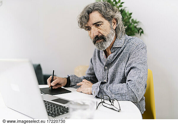 Senior male business professional looking at laptop while using graphics tablet at home office