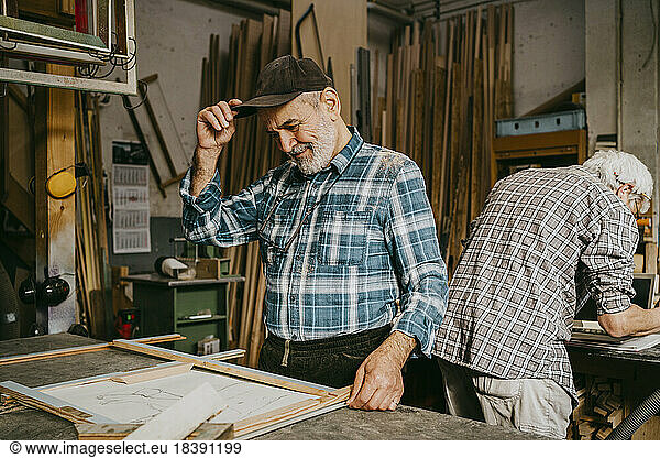 Senior male carpenter adjusting cap while working with colleague at repair shop