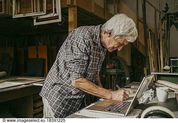 Senior male carpenter preparing document with laptop on workbench at repair shop