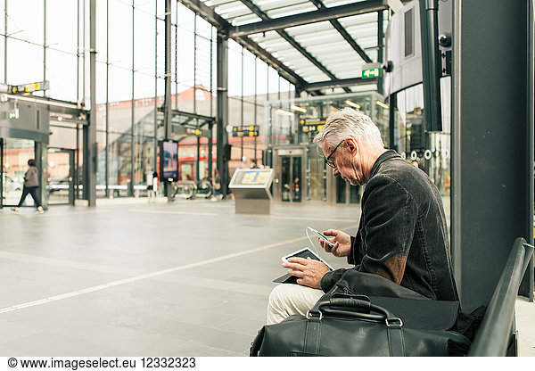 Senior male commuter sitting with technologies and bags at railroad station