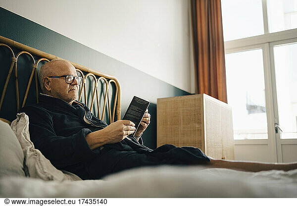 Senior male reading book while sitting on bed in bedroom