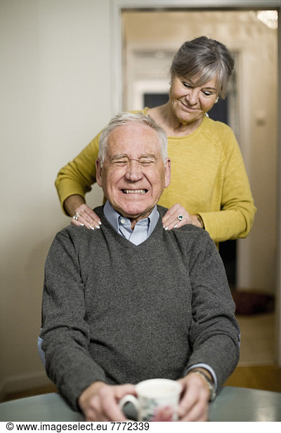 Senior man clenching teeth while woman giving him a shoulders massage at home