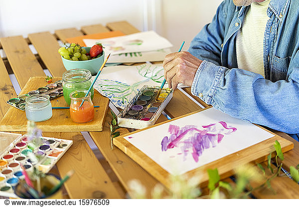 Senior man dipping paintbrush in watercolor paints on table at home