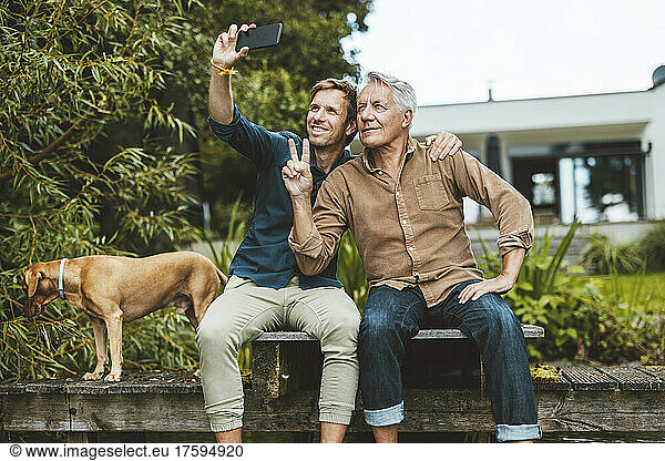 Senior man gesturing peace sign taking selfie with son through mobile phone at backyard