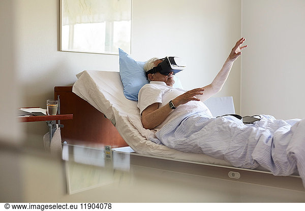 Senior man gesturing while using VR glasses on bed in hospital ward