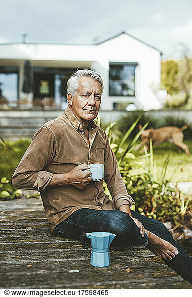 Senior man holding coffee cup sitting on jetty at backyard