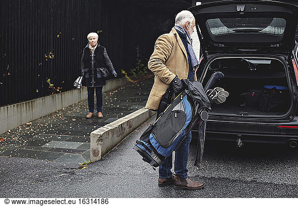 Senior man holding golf bag by car trunk while female partner standing on sidewalk during winter