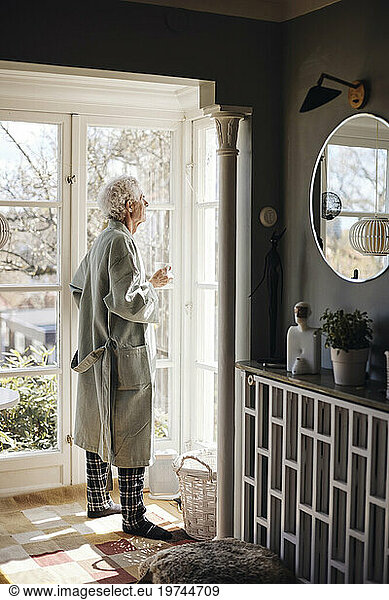 Senior man in bathrobe holding coffee cup while looking outside window at home