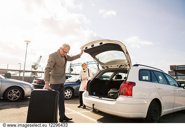 Senior man loading luggage in car trunk with woman standing in parking lot against sky