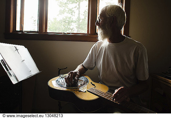 Senior man looking at papers while learning guitar against window at home