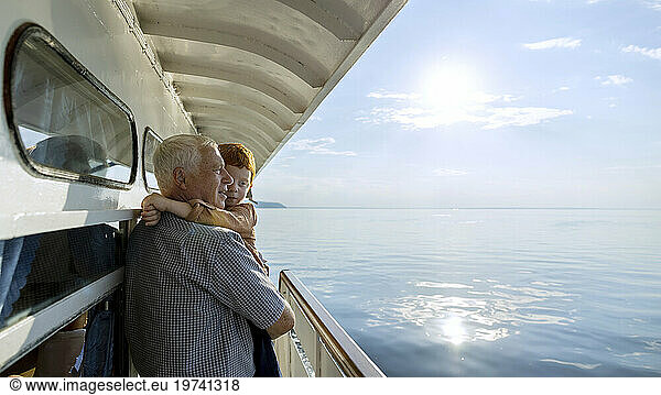 Senior man looking at sea from ship and carrying grandson on sunny day