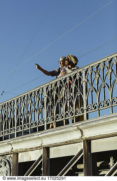 Senior man pointing while standing by woman on bridge against blue sky