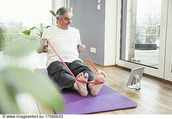 Senior man pulling resistance band watching online tutorial through tablet PC at home