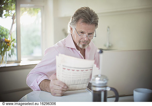 Senior man reading newspaper at laptop at morning kitchen table