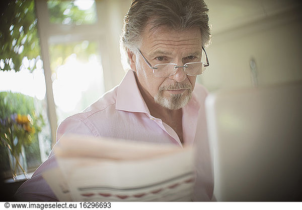 Senior man reading newspaper at laptop in sunny morning kitchen