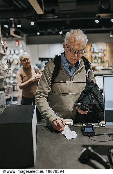 Senior man signing bill by credit card reader at checkout counter in appliances store