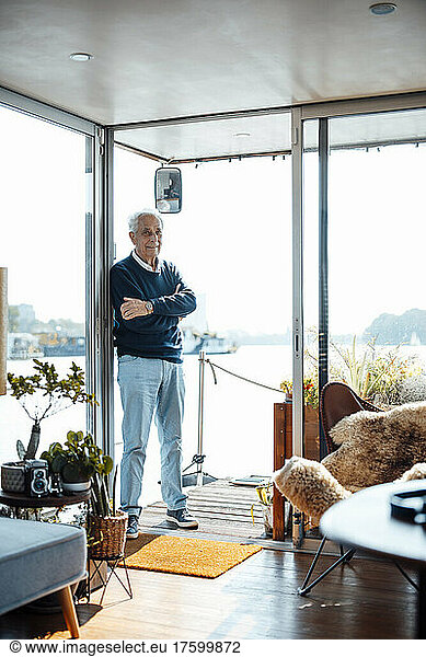Senior man standing with arms crossed at houseboat doorway