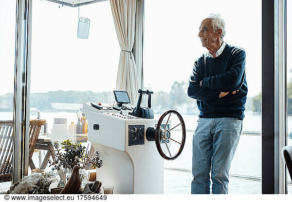 Senior man standing with arms crossed by window at houseboat