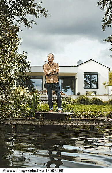 Senior man standing with arms crossed on jetty at backyard