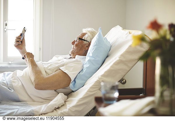Senior man taking selfie with smart phone on bed in hospital ward