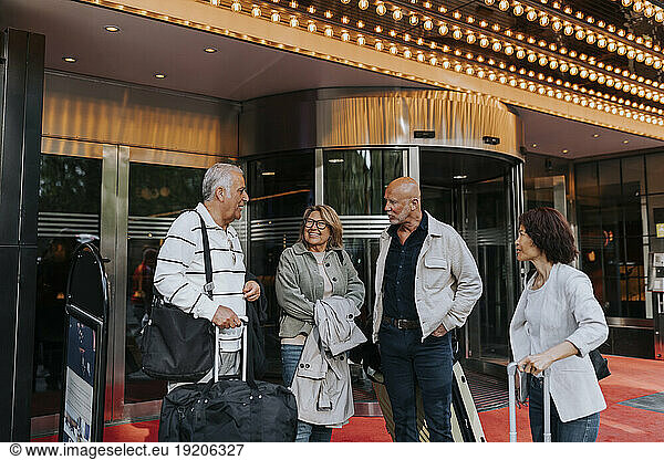 Senior man talking to male and female friends standing with luggage outside movie theater