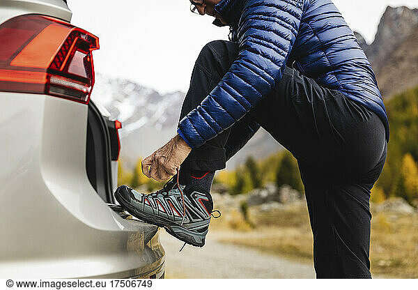Senior man tying lace of hiking boot at car trunk on vacation