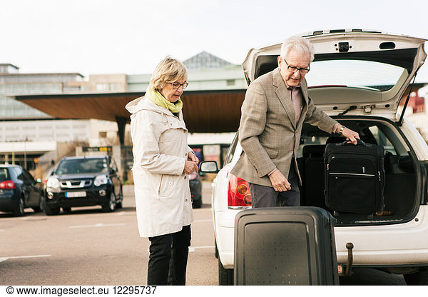 Senior man unloading luggage from car trunk by woman standing in parking lot