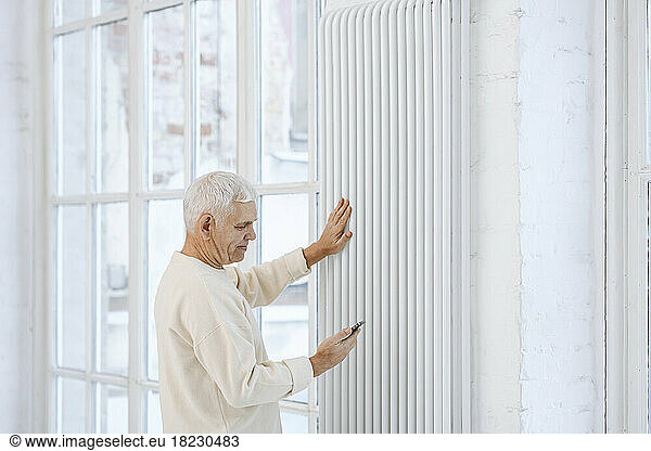 Senior man using smart phone touching radiator at home