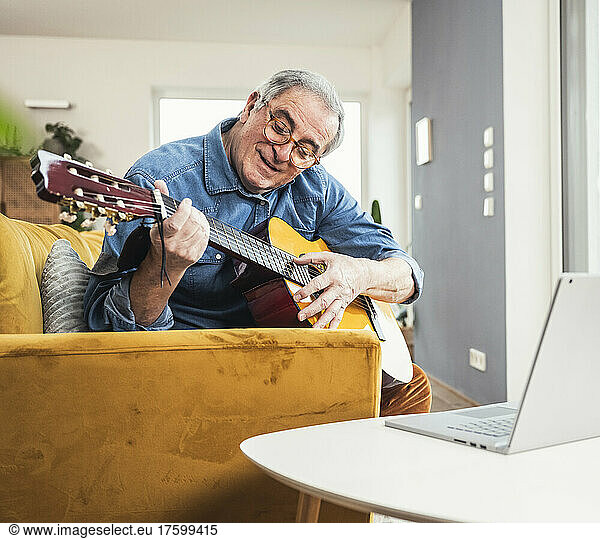 Senior man wearing eyeglasses playing guitar on sofa in living room