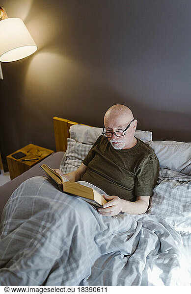 Senior man wearing eyeglasses while reading book on bed at home