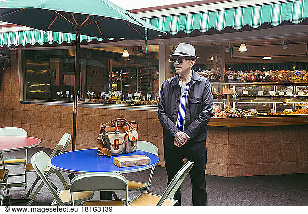 Senior man wearing hat standing by table at sidewalk cafe