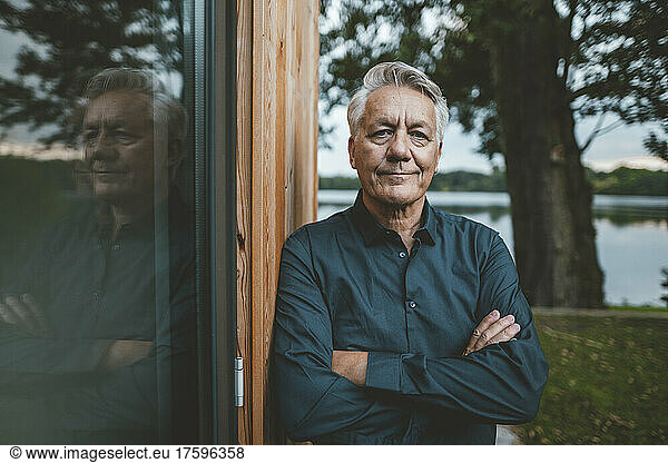 Senior man with arms crossed standing by glass wall at backyard