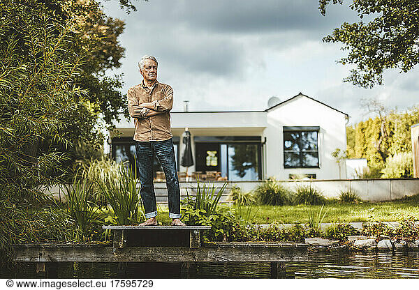 Senior man with arms crossed standing on jetty by lake at backyard