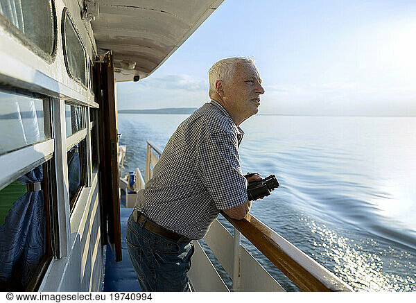 Senior man with binoculars in hand looking at sea from ship on sunny day