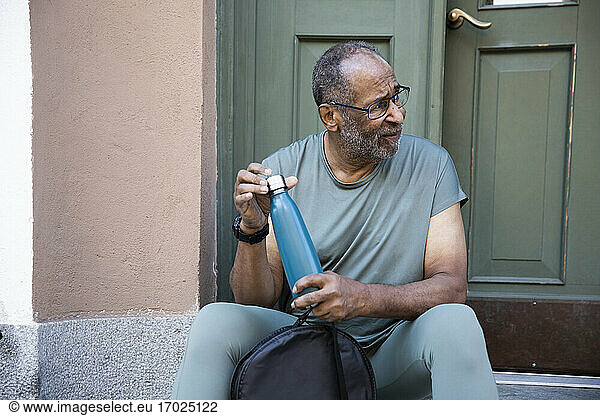 Senior man with bottle looking away while sitting against doorway