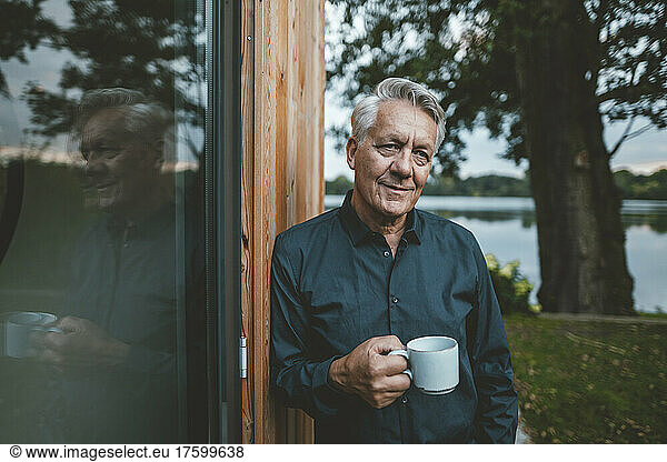 Senior man with coffee cup standing by glass wall at backyard