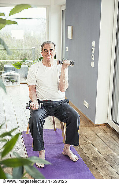 Senior man with dumbbells sitting on chair working out at home