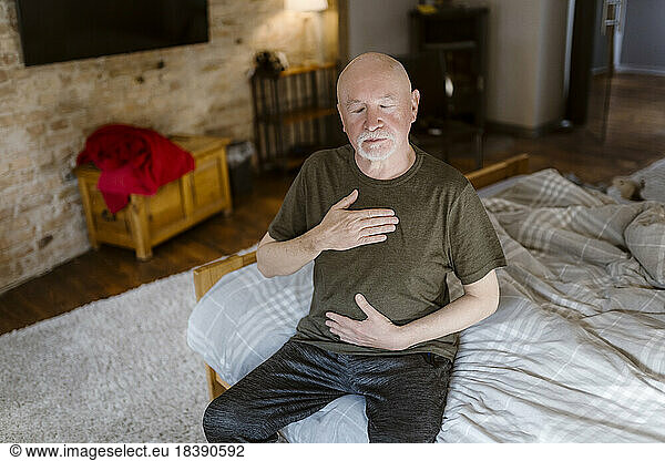 Senior man with eyes closed doing breathing exercise while sitting on bed at home